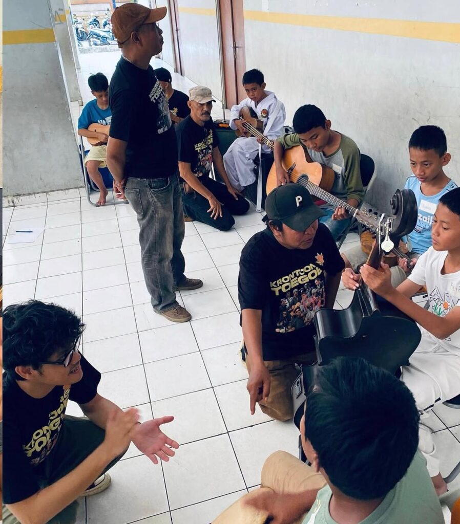 Whatsapp image 20260612 at 19 57 57 catrawarta A man standing and guiding a guitar lesson outdoors among several kids seated with guitars in a tiled courtyard